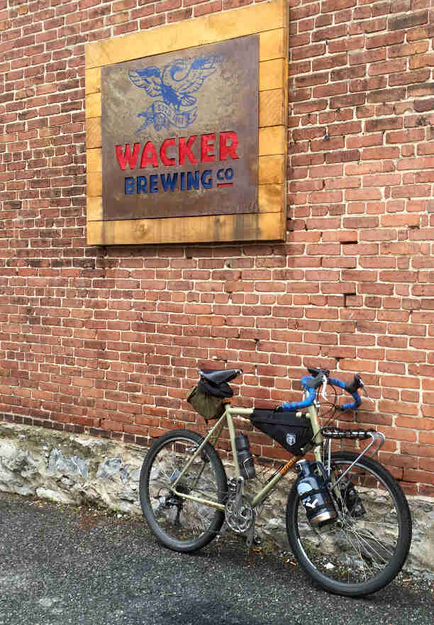 Right side view of a bike with beer growlers strapped to the fork, parked along a brick wall with a Wacker Brewing sign