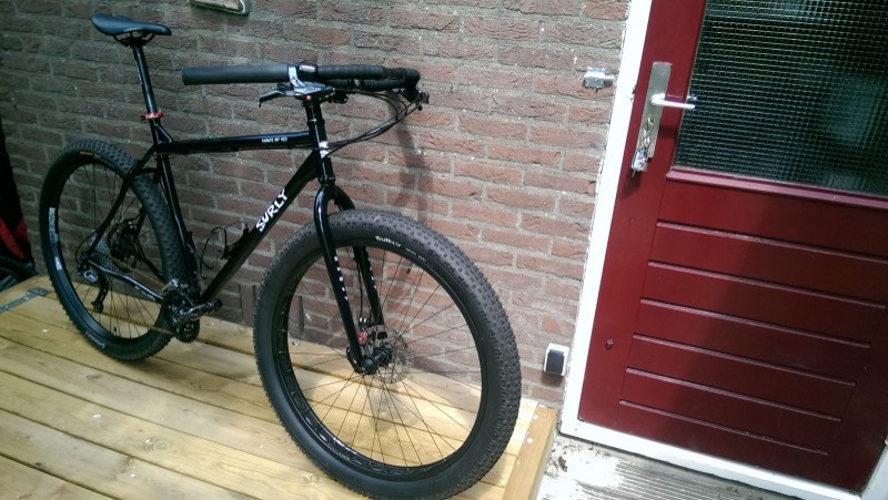 Right side view of a black Surly bike, on a wood deck, leaning on a brick wall next to a red door