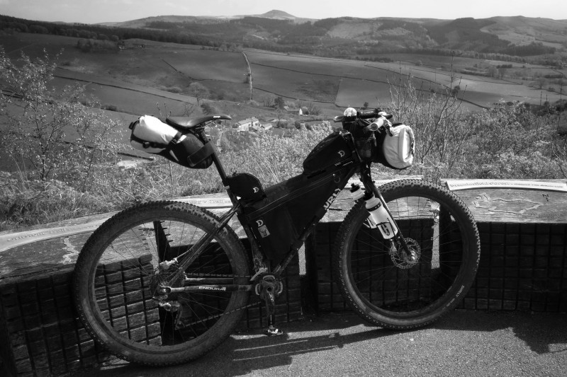 Right side view of a Surly bike with gear packs, leaning on a short stone wall, with hilly farm fields behind it