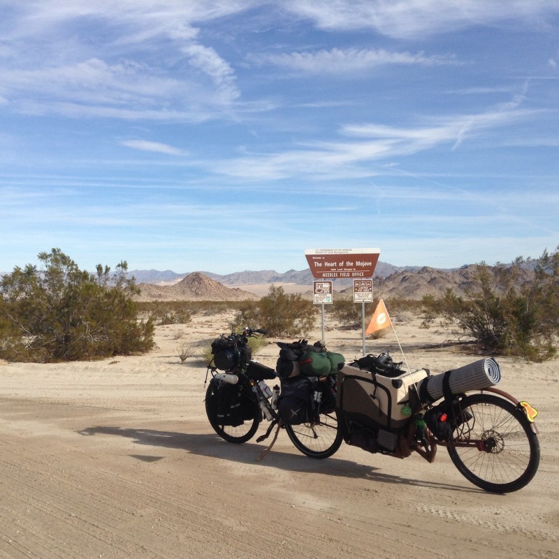Left side view of a Surly bike and trailer, both loaded with gear, on a sandy road in the Mojave desert