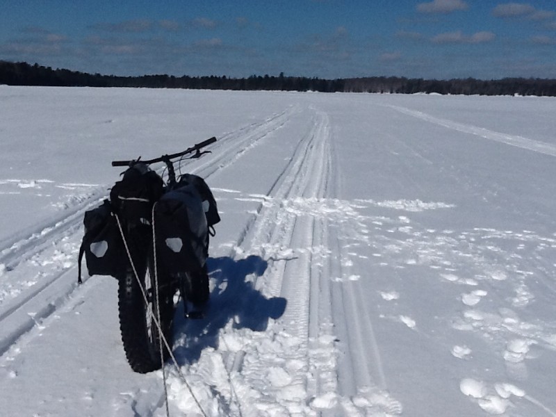 Right view of a Surly fat bike with saddlebags, parked on a snowy frozen lake, with trees in the background