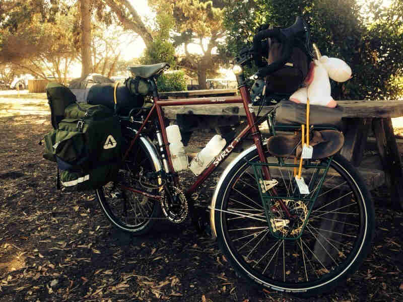 Right side view of a red Surly Disc Trucker bike with gear, against a picnic table, in an area with mulch covered ground