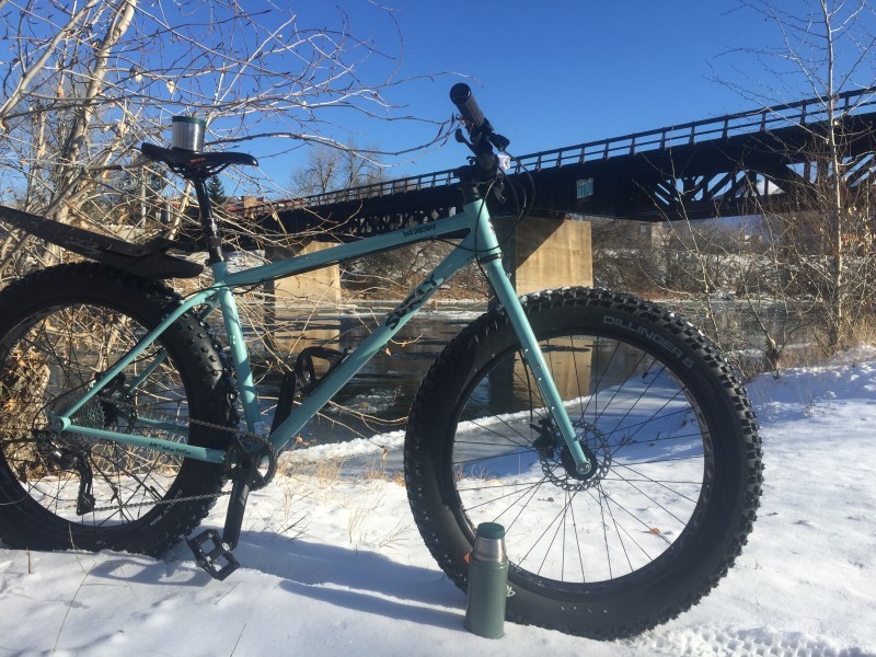 Right side view of a mint Surly Wednesday fat bike, parked on snow, next to a river, with a bridge in the background