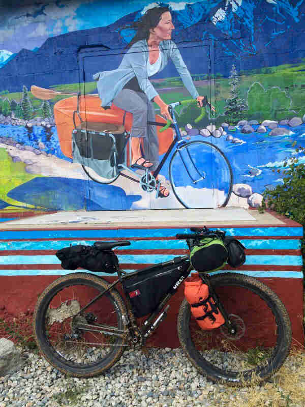 Right side view of a muddy, black Surly fat loaded with gear packs, parked in front of a wall with a mural