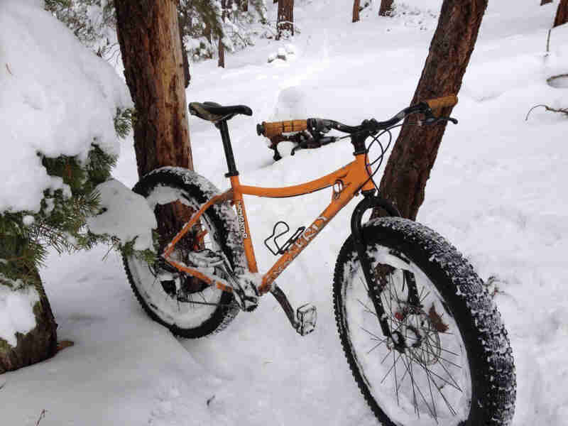 Right side view of an orange Surly Instigator fat bike, parked in deep snow next to trees