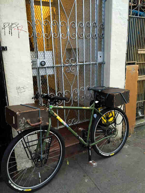Left side view of green bike with boxes on the front and rear racks, parked in front of a gated door