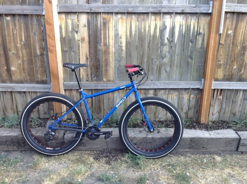 Right side view of a blue Surly Pugsley fat bike, parked along a railroad tie, on a yard, in front of a wood fence wall