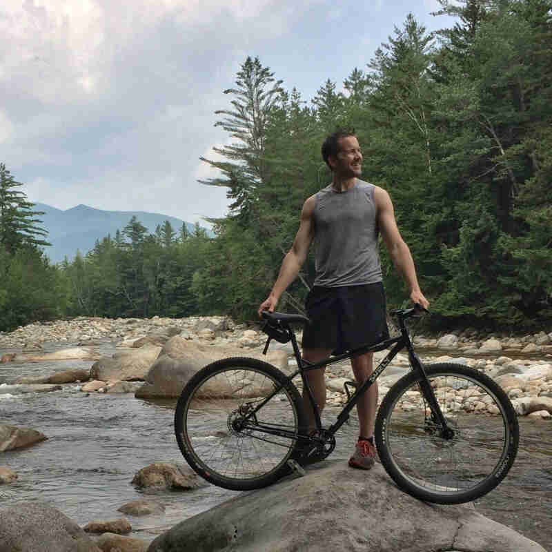 Right side view of a black Surly Karate Monkey bike, with a cyclist behind, on a rock next to river in the mountains