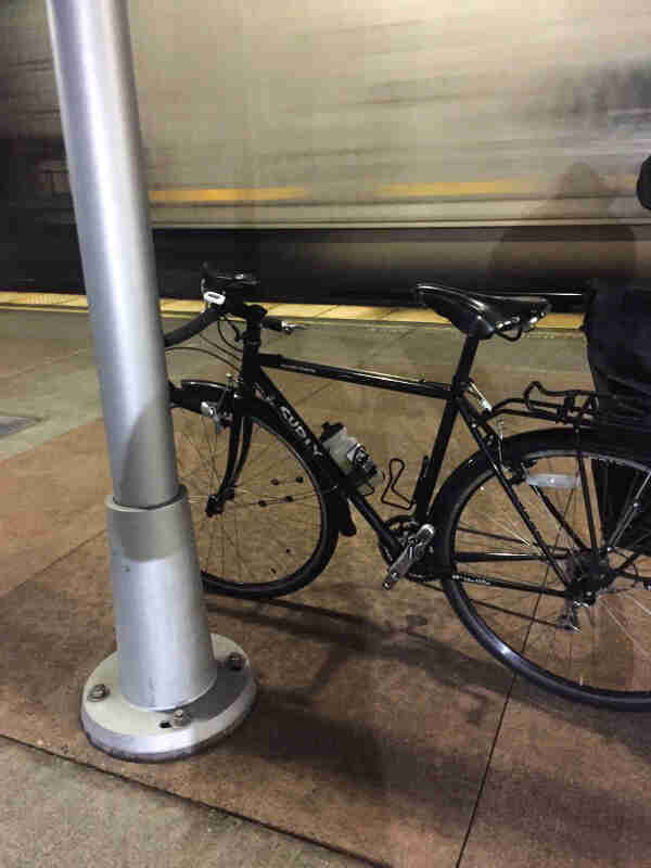 Left side view of a black Surly bike, parked on subway platform next to a steel pole