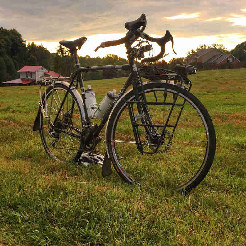 Right side view of a Surly bike, with racks, parked in grass field, with 2 houses in the background, at sunrise