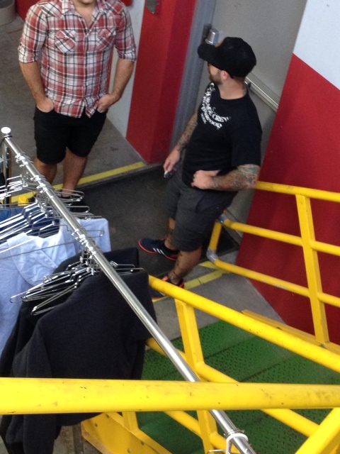 Downward view of a person with tattooed arms, standing at the bottom of a staircase, talking to another person