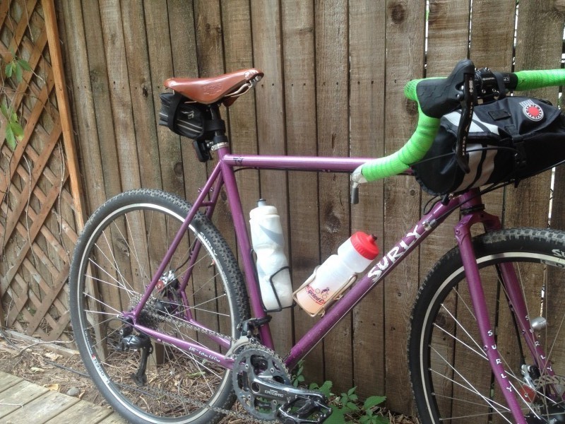 Right side view of a purple Surly bike with water bottles and handlebar pack, leaning against a wood fence wall