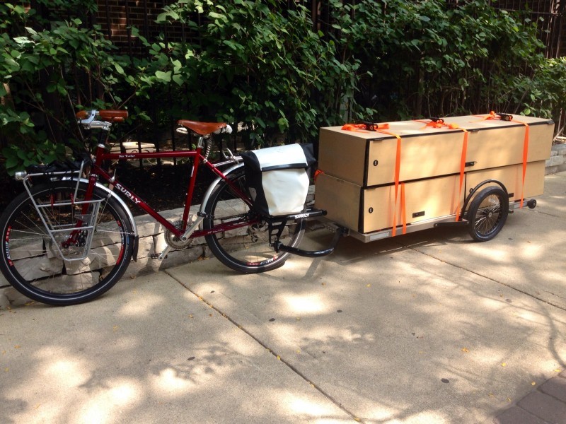 Left side view of a red Surly Disc Trucker bike with a trailer loaded with cabinets behind, parked on a sidewalk