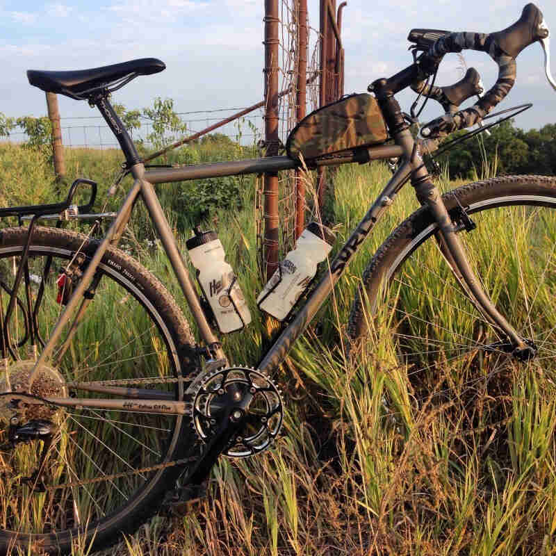 Right side view of an olive drab Surly Cross Check bike, parked in tall grass, leaning against the side of a wire fence