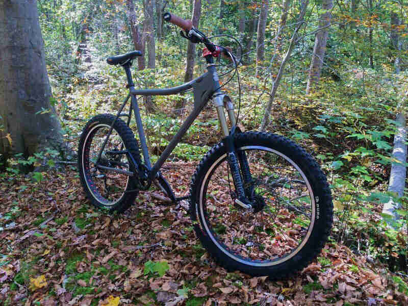 Front right view of an olive green Surly Instigator fat bike, standing on leaves, in a thick forest