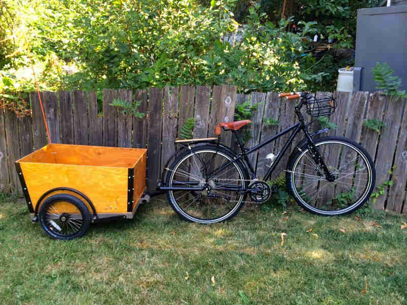 Right side view of a Surly bike, black, with a trailer hitched on back, parked in a grass yard against a wood fence