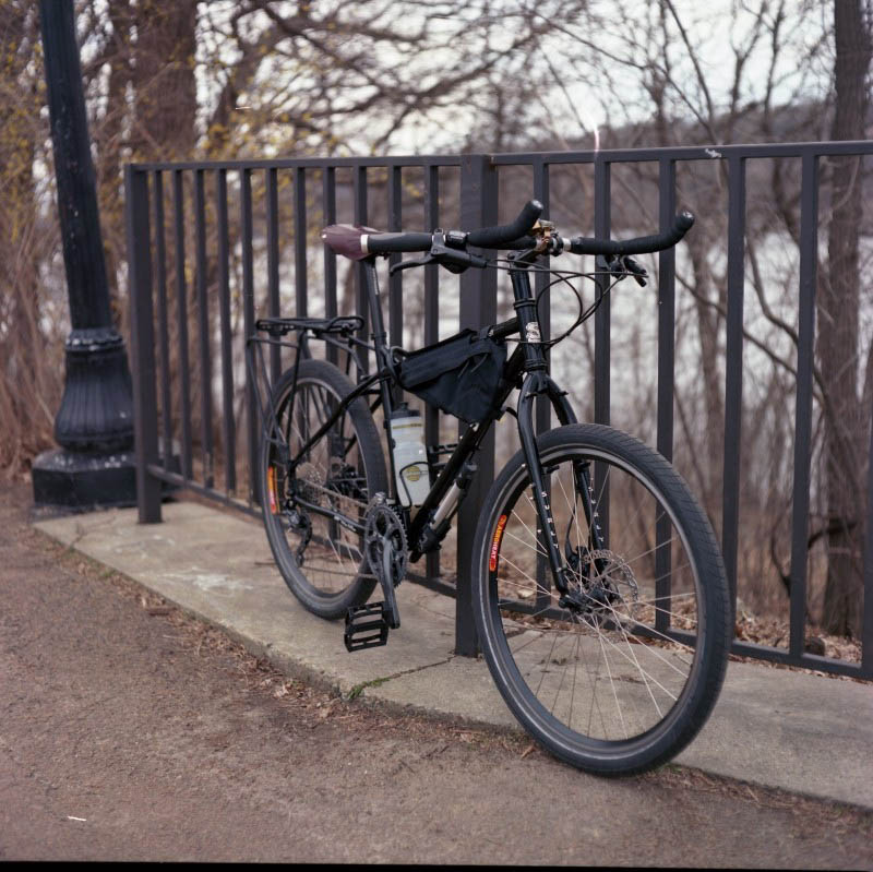 Front right side view of a Surly bike, black, parked on concrete alongside a iron fence, with trees in the background