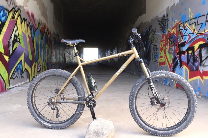 Right side view of a tan Surly Instigator bike, parked at the entry of a concrete tunnel with graffiti on the side walls