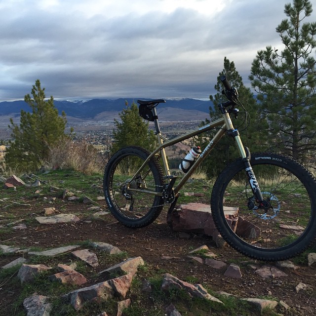 Right side view of a Surly Instigator bike, parked on a rocky hilltop clearing, overlooking a town in the foothills