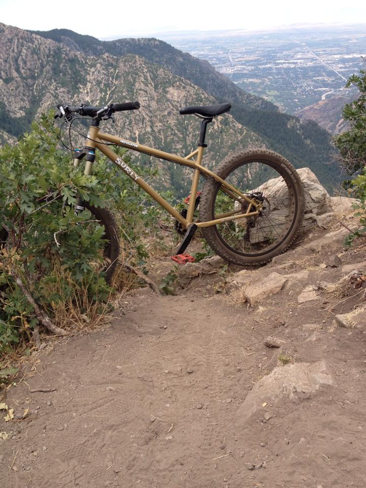 Left side view of a Surly Instigator bike, parked on rocks at the edge of a cliff, with a mountain valley in background