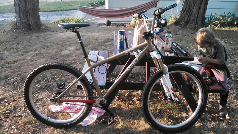 Right side view of a tan Surly Instigator bike, in a yard, leaning against a picnic table with a child sitting at it