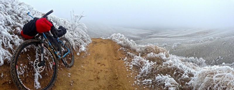 Front, left side view of a blue Surly bike with gear, parked on a dirt trail, in a frosted, grass field with hills
