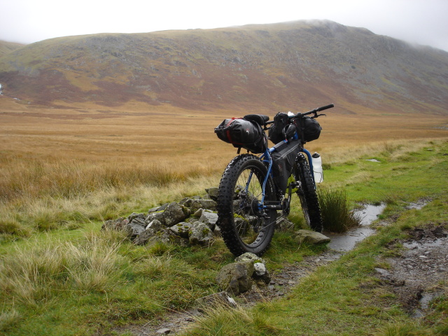 Rear, right side view of a blue Surly fat bike with gear, next to a pile of rocks, facing a field with mountains behind