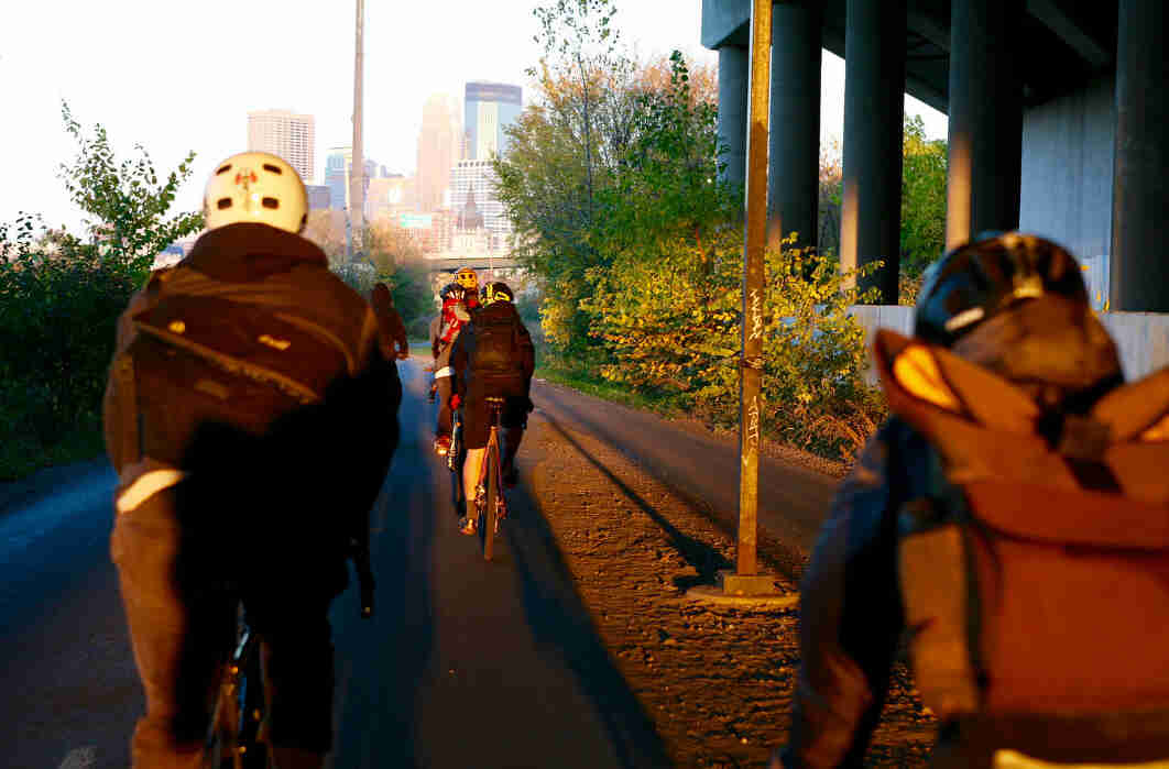 Rear view of a group of cyclists riding under a bridge towards a city skyline in front of them