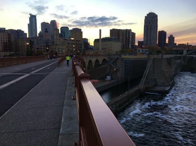 A view straight down the sidewalk of a bike bridge over a river, with a city skyline ahead