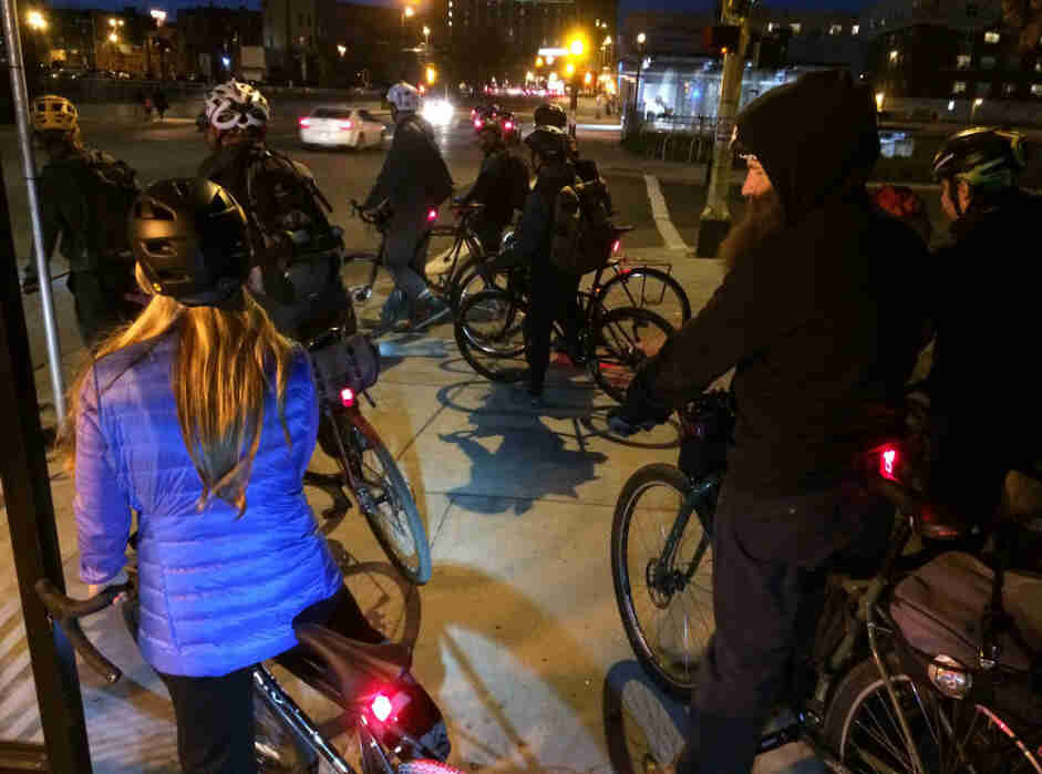 Rear view of a group of cyclists standing with their bikes on a city street corner at night