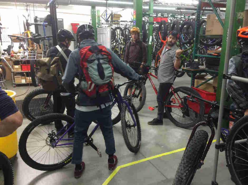 A group of cyclists standing with bikes in warehouse, with hanging bikes in the background