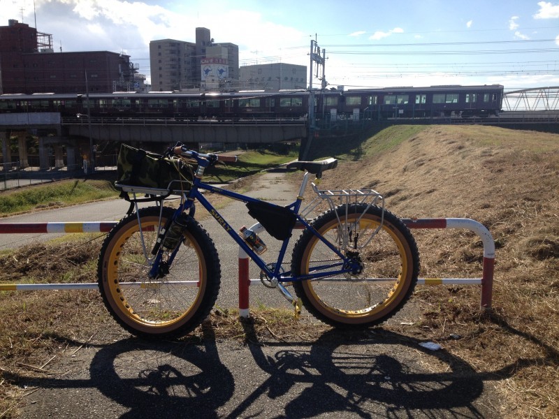 Left side view of a blue Surly Pugsley bike, parked across a paved trail, with rail cars on a bridge in the background