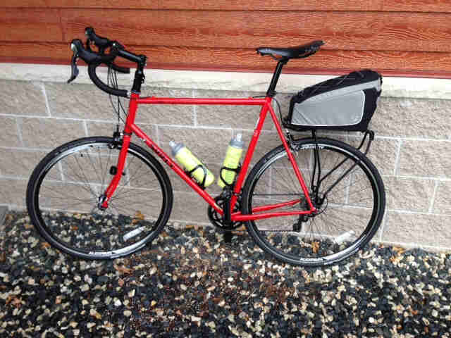 Left side view of a red Surly Pacer bike with a rear rack pack, parked in rocks next to a stone block and wood wall