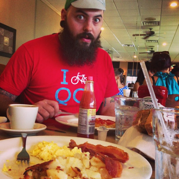 Front view of a person with a beard, sitting behind a table with breakfast foods, inside a restaurant