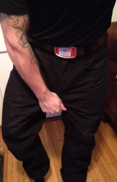 Front, chest down view of a person wearing black Surly pants, pulling on the crotch fabric, in a room with wood floors
