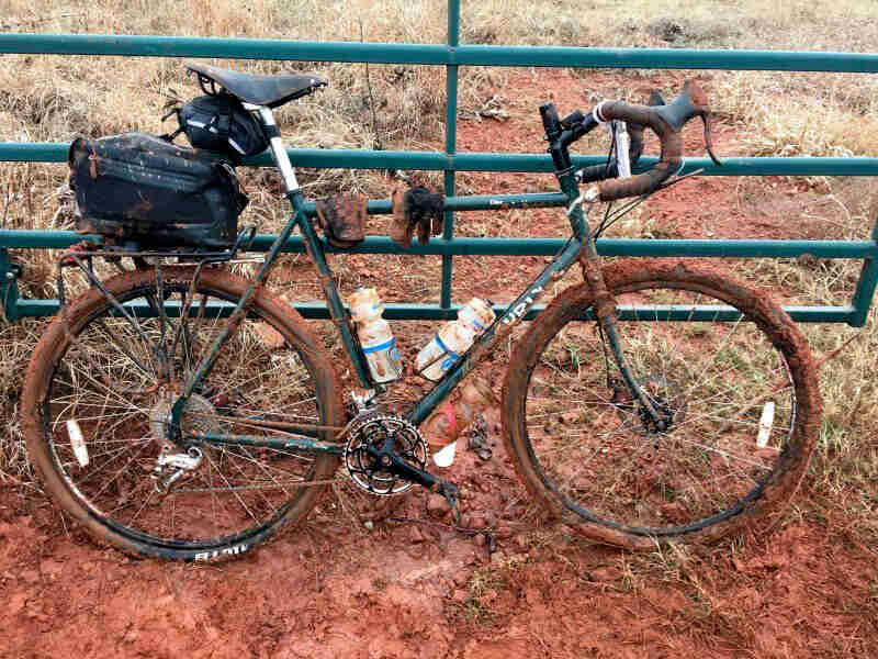 Right side view of a muddy, green Surly bike, parked in red mud in front of a gate