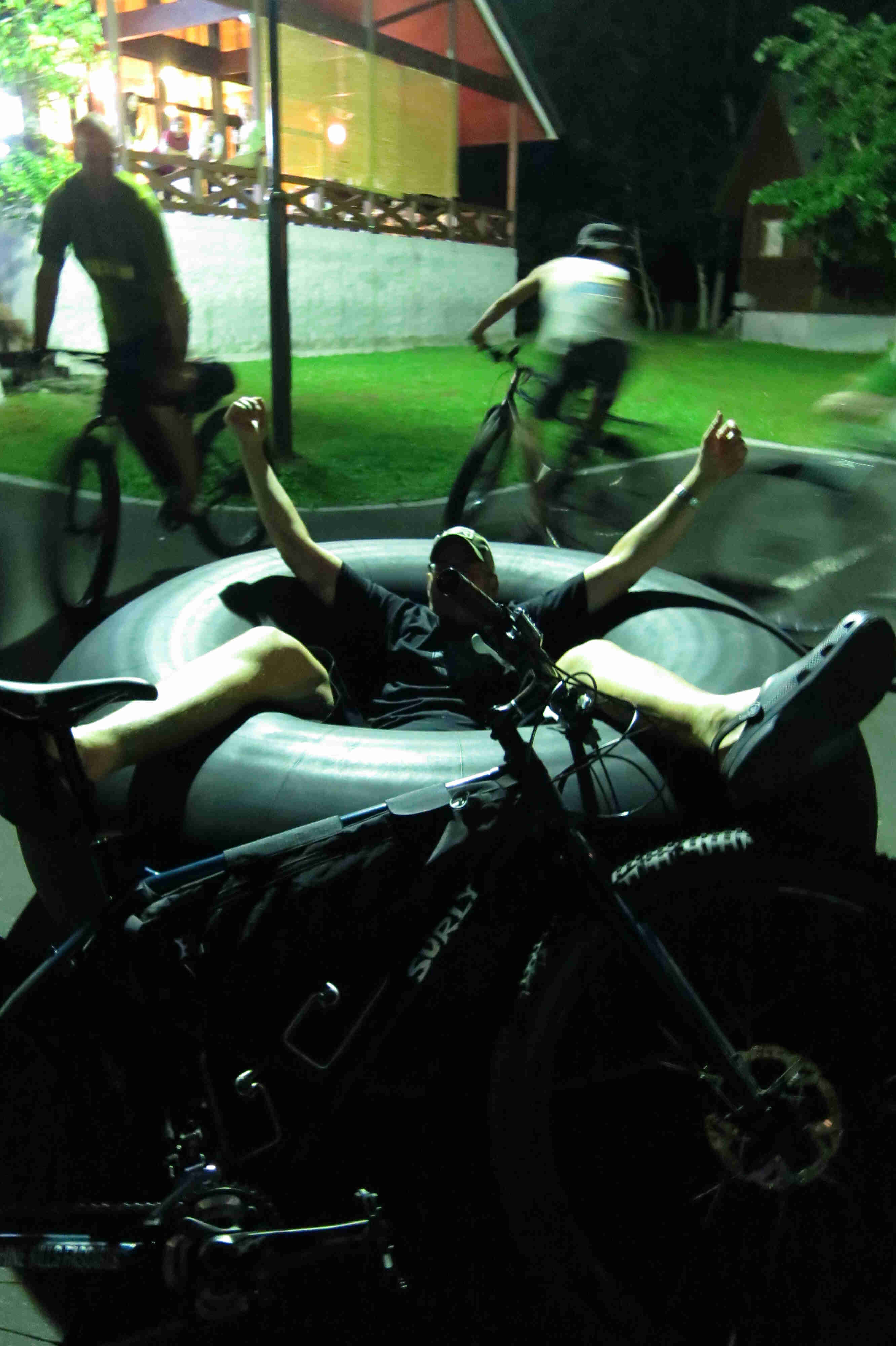 Right side view of Surly fat bike, leaning on a large inner tube with a person laying in it, on a parking lot at night 