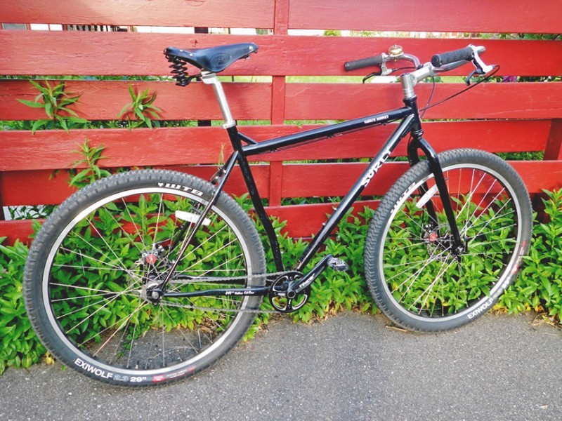 Right side view of a black Surly Karate Monkey bike, leaning against a wood fence wall, with weeds behind the wheels