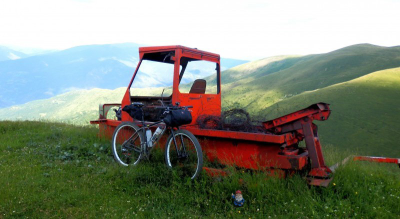 Right side view of a bike with gear, parked against a red, broke down piece of machinery, in the grassy, rolling hills