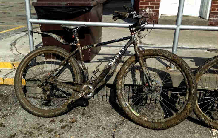 Right side view of a black Surly Karate Monkey bike, covered in mud, parked on a paved lot and leaning on a metal rail 