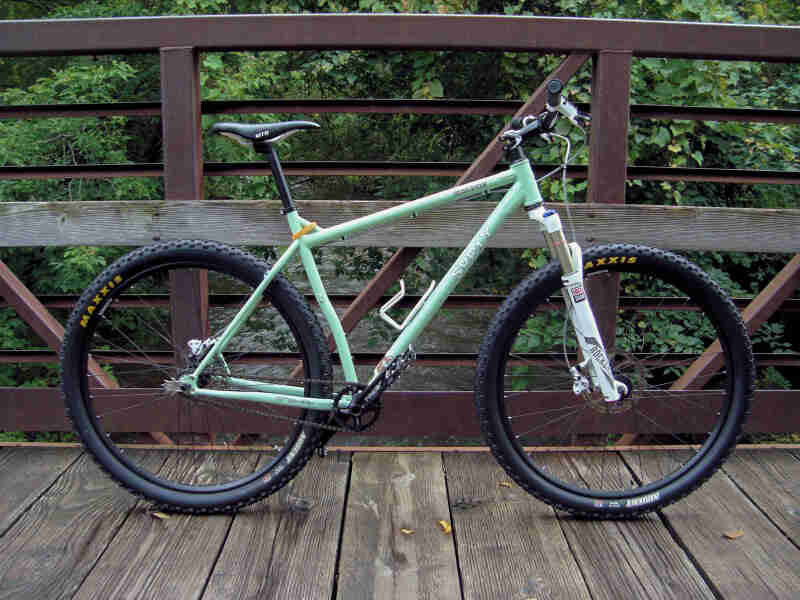 Right side view of a mint Surly Karate Monkey bike, parked on a bridge above a stream with trees on the sides