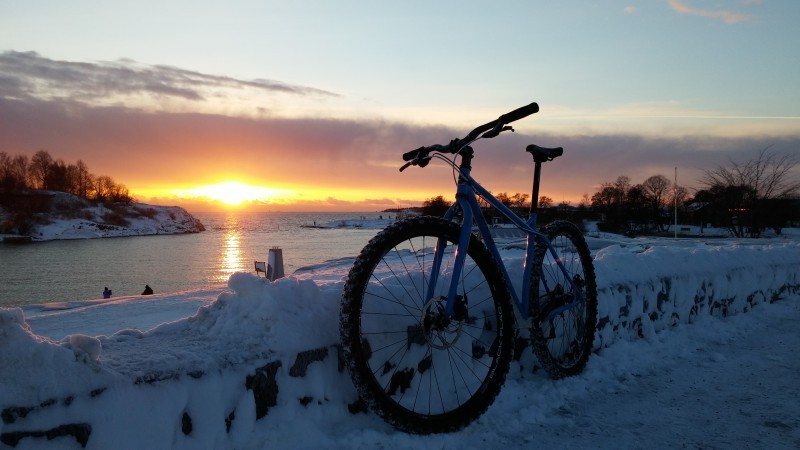 Left side view of a blue Surly Karate Monkey bike, leaning on a short snowy wall along a river at sunset