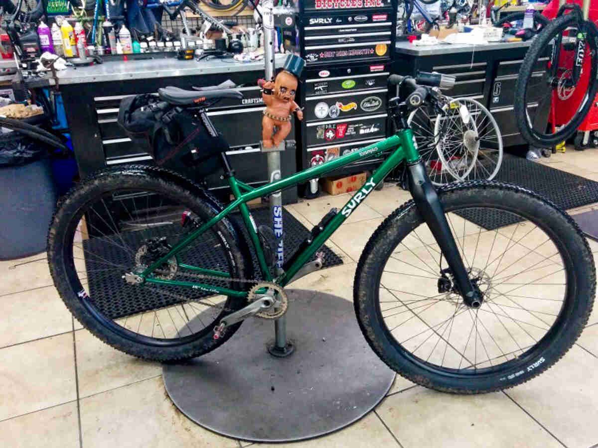 Right side view of a green Surly Krampus bike, parked against a pole with a doll on it, in a bike shop