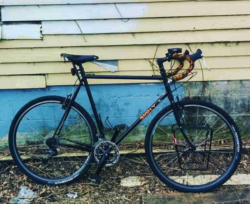 Right side view of a black Surly Cross Check bike, parked in mulch, next to the wall of a house