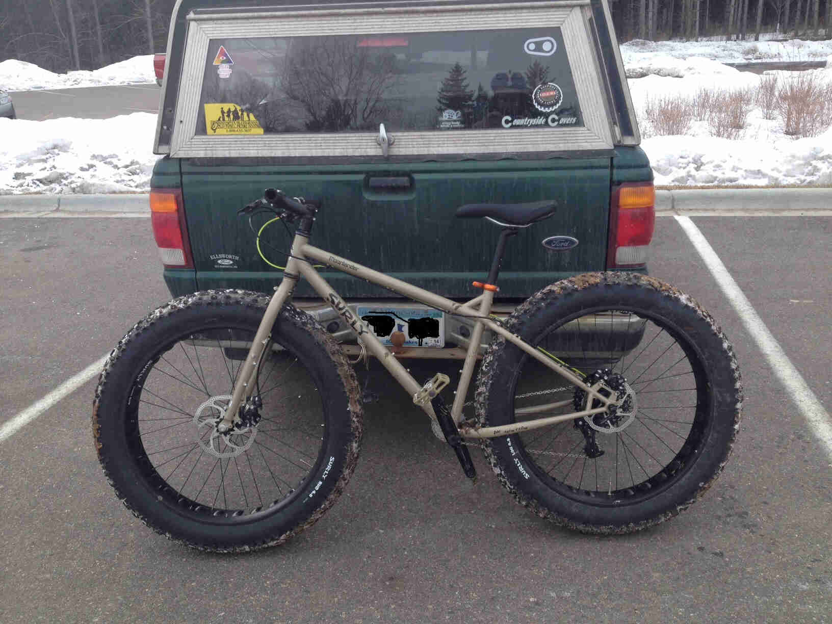 Left side view of a tan Surly Moonlander fat bike, leaning against the tailgate of a pickup, in a paved parking lot spot