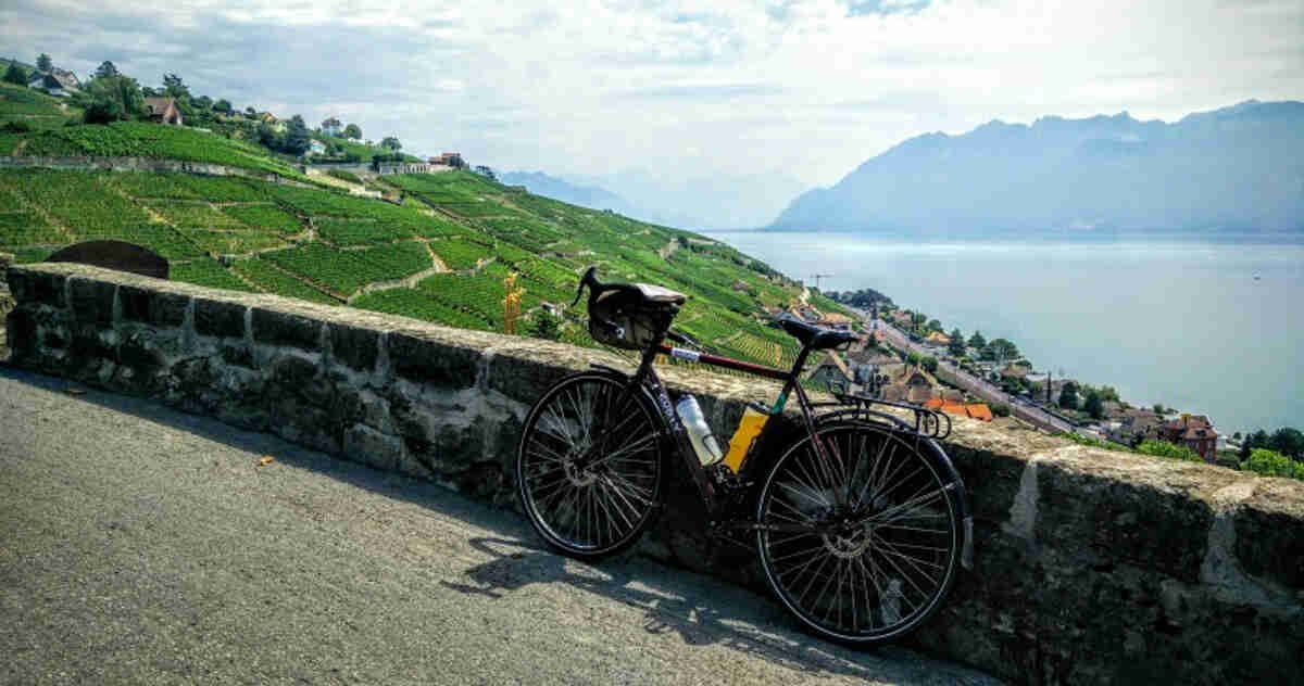 Left side view of a Surly bike parked against a short stone wall, with a green hill, a lake and mountains behind