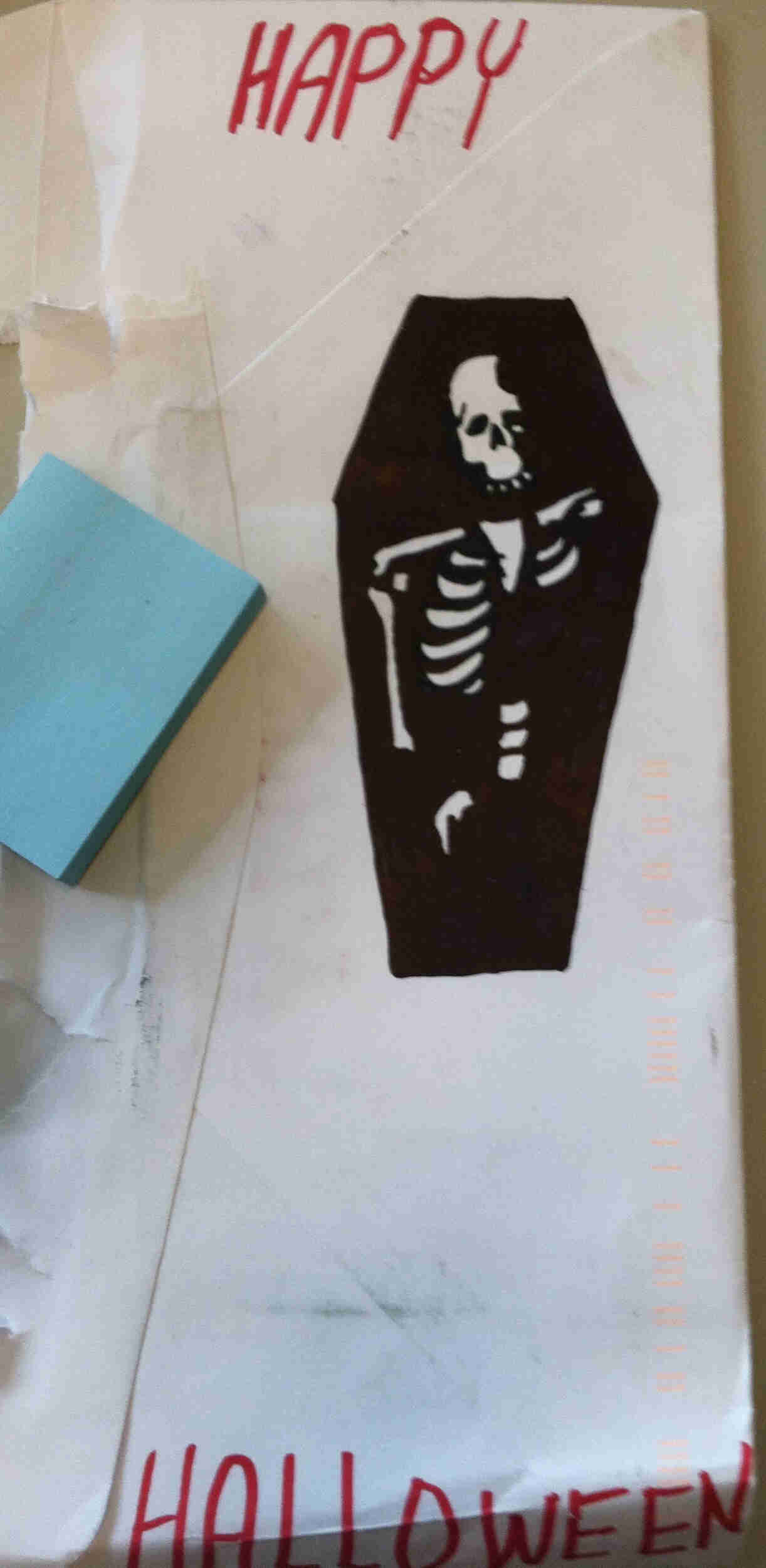Downward, view of the back of a white envelope, with Happy Halloween, written in red marker, and a coffin drawing on it