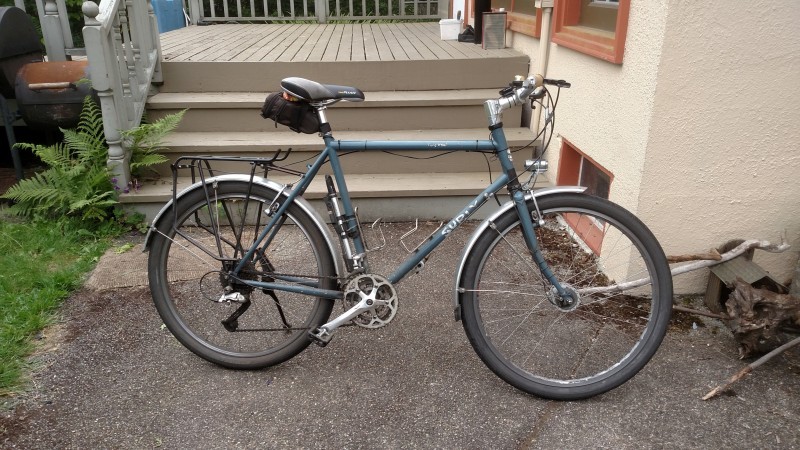 Right side view of a gray Surly Long Haul Trucker bike, parked on pavement in front of the stairs on a house deck