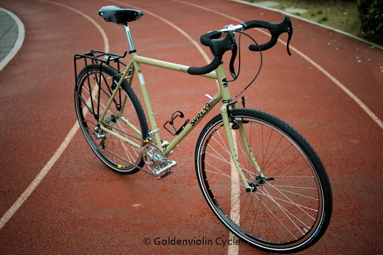 Right side view of a light green Surly Long Haul Trucker bike, parked across a running track, near one of the turns