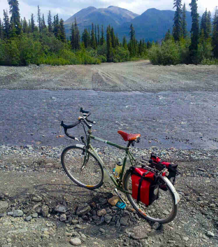 Downward, left side view of a green Surly Long Haul Trucker bike, on a rock river bank, with mountains in the background